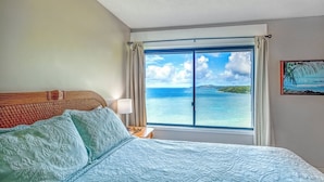 A hotel room with a bed, a nightstand, a window with a view of the ocean, and a painting on the wall.