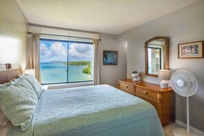 A hotel room with a large bed, a dresser, a standing fan, a mirror, and a view of the ocean. - Charming & Affordable Couple's Retreat w/ Ocean Views from Every Room (Princeville)