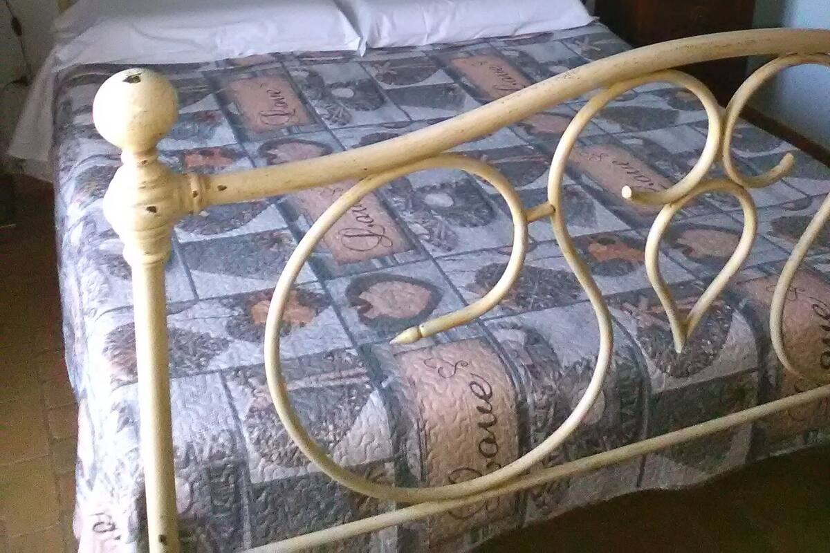 Una cama con estructura metálica y colcha estampada. Se observa una mesita de noche con una lámpara.