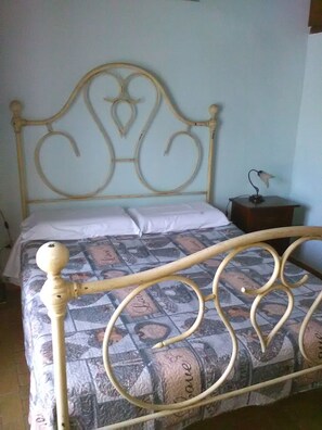 Ein Bett mit Metallgestell und einem gemusterten Bettbezug. Ein Nachttisch mit einer Lampe steht daneben.