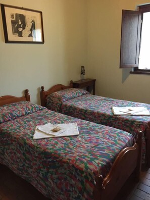 Ein Zimmer mit zwei Holzbetten, jeweils mit einem farbenfrohen, gemusterten Überwurf. An der Wand hängt ein gerahmtes Bild, ein kleiner Tisch mit einer Lampe steht bereit, und ein Fenster mit einer Holzjalousie.