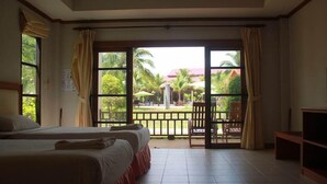 Ein Hotelzimmer mit zwei Betten, einem Balkon mit Blick auf Palmen und einen Pool, sowie einem Fernseher auf der rechten Seite.