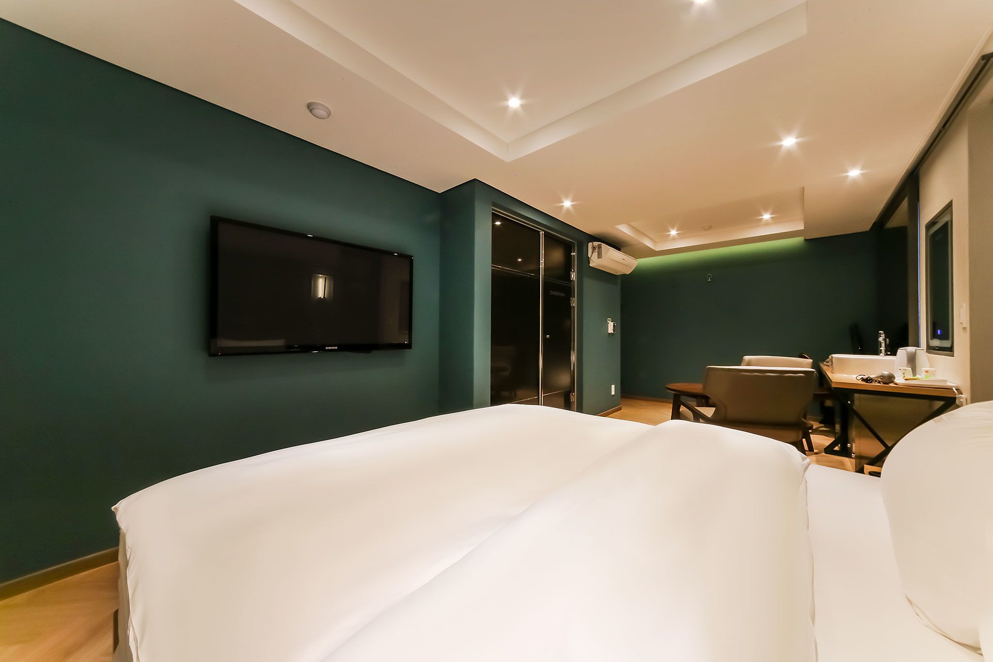 A hotel room with a bed, a TV mounted on the wall, a desk with a chair, and a window.