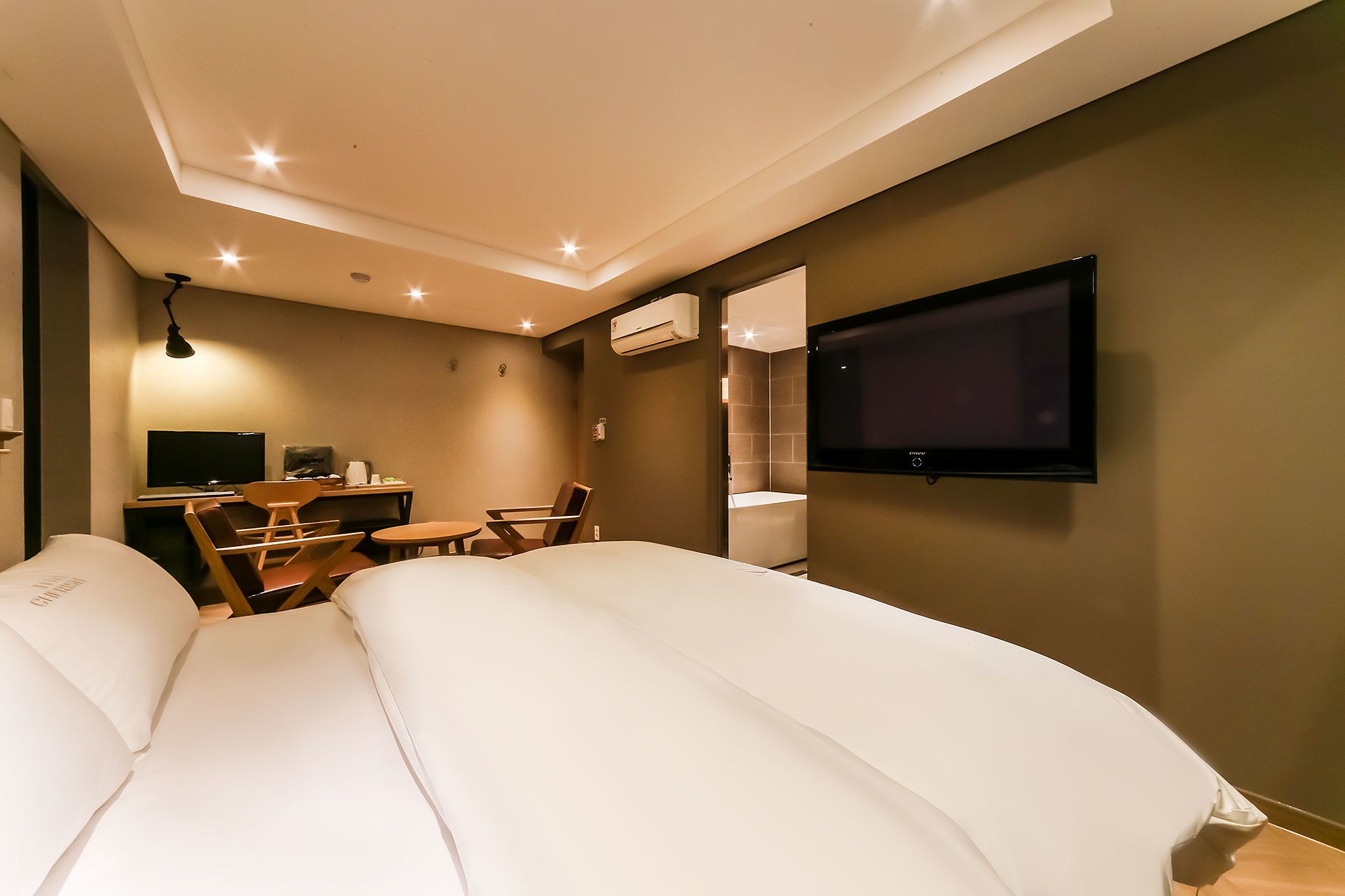 A modern hotel room with a large bed, a desk with a chair, a TV mounted on the wall, and an air conditioning unit.