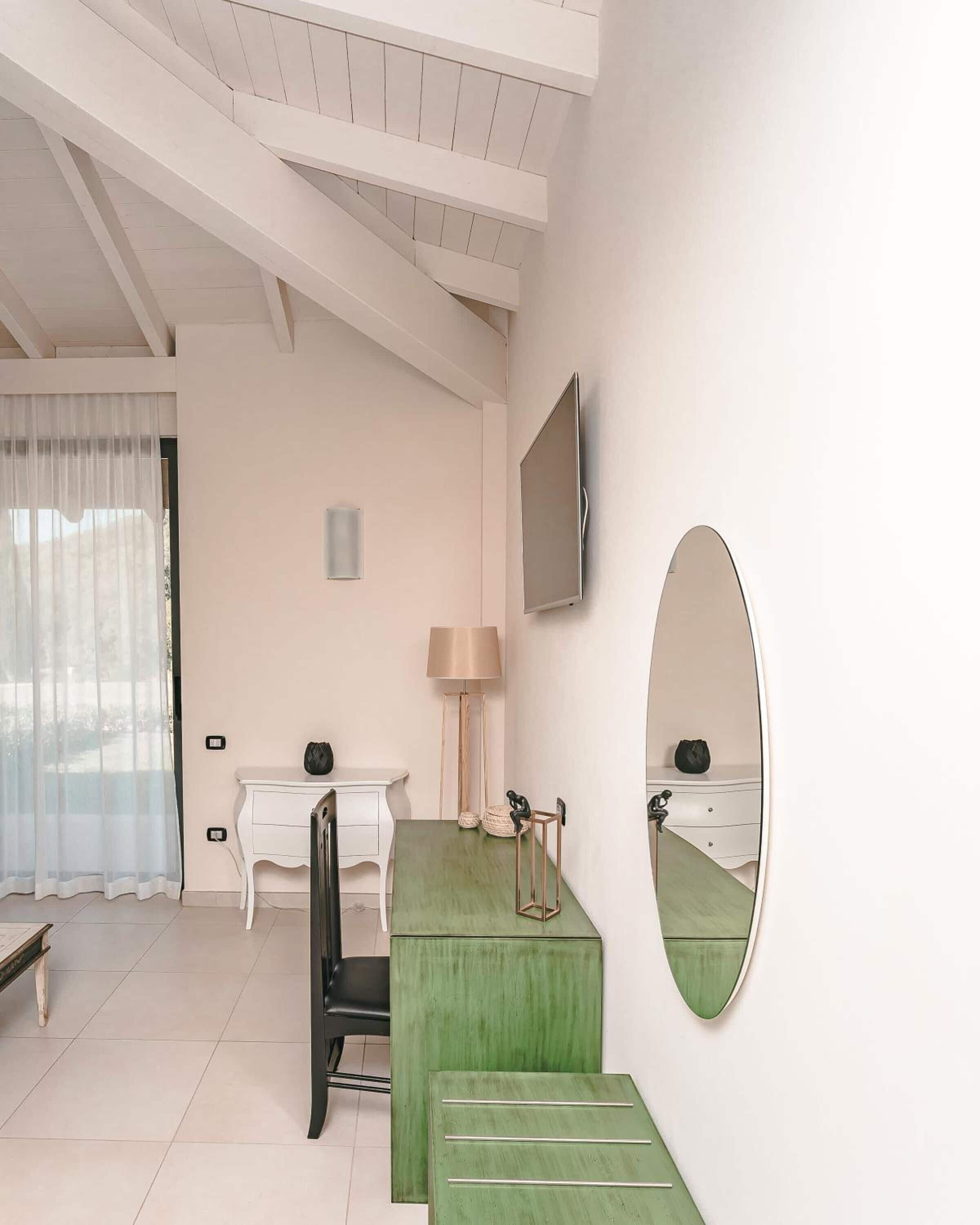 A modern bedroom with a green vanity, a large mirror, a flat-screen TV, and a window with sheer curtains.