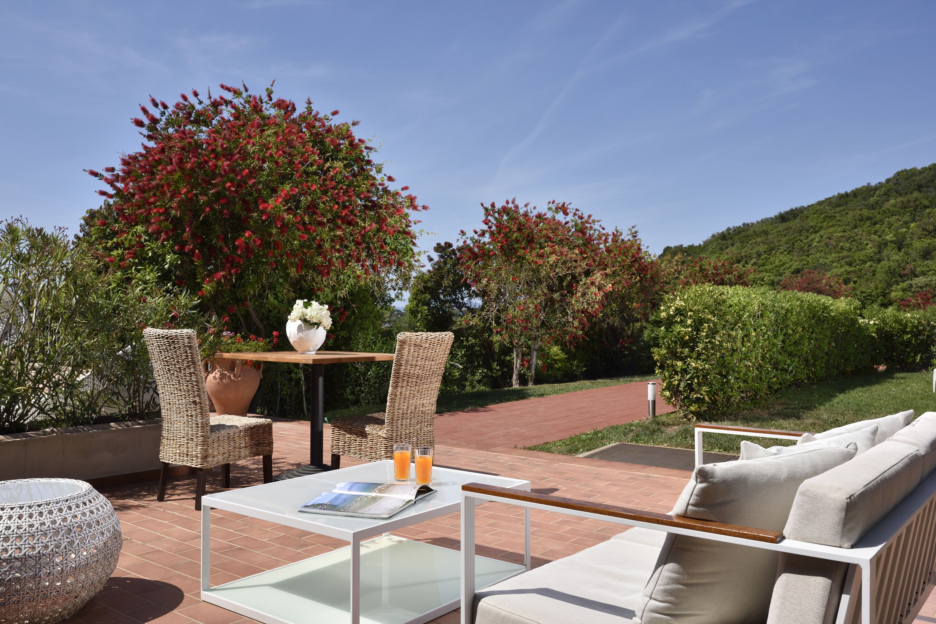An outdoor patio with wicker furniture, a glass-top table, and a view of a landscaped garden with red flowering bushes.