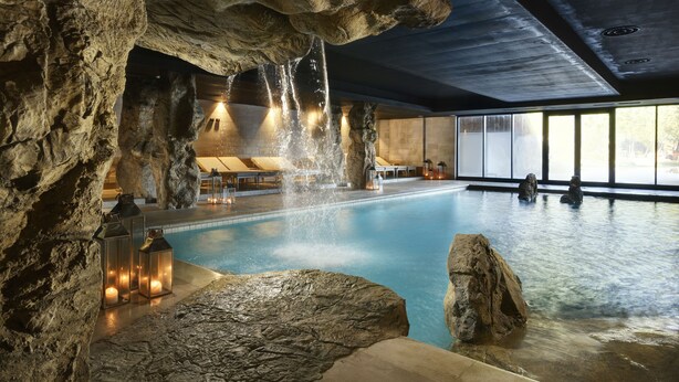 Una piscina con formazioni rocciose naturali, illuminazione calda e vista sull'esterno attraverso ampie finestre.