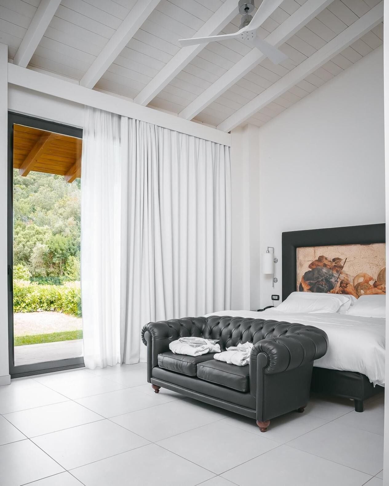 A modern bedroom with a black leather sofa, a bed with white linens, and a large window with white curtains.