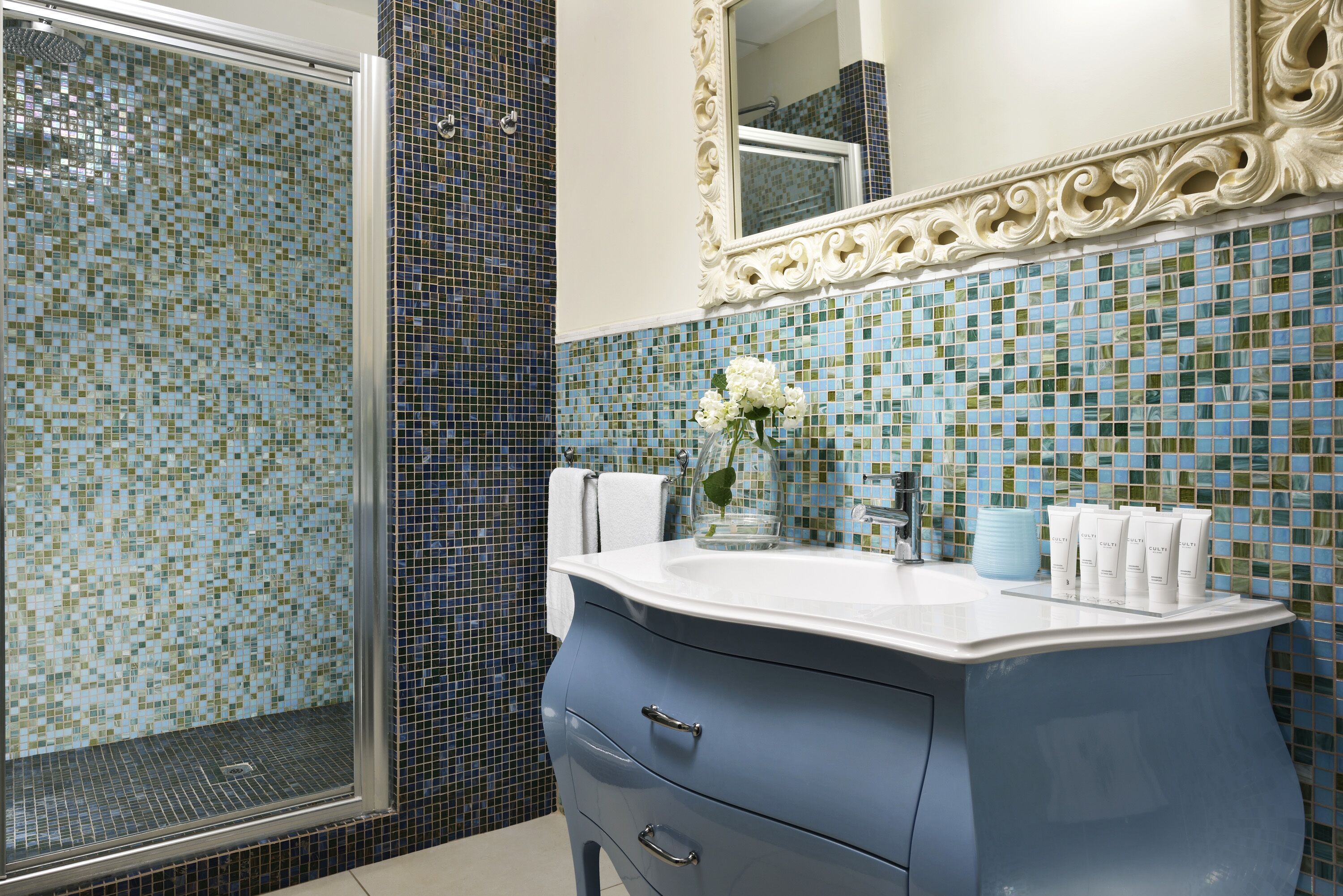 Un baño con ducha, un lavabo con artículos de aseo y un espejo con marco decorado.