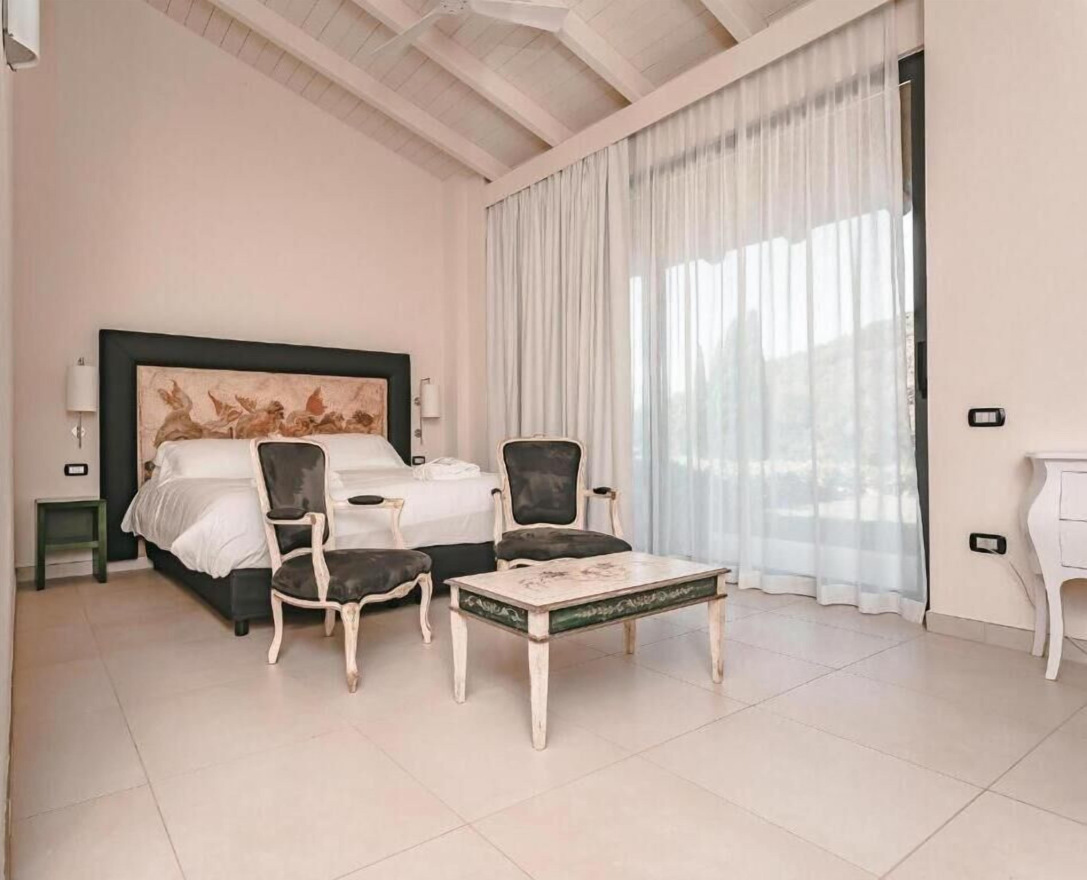 Habitación de hotel con una cama grande, dos sillones, una mesita y vistas al exterior a través de cortinas translúcidas.