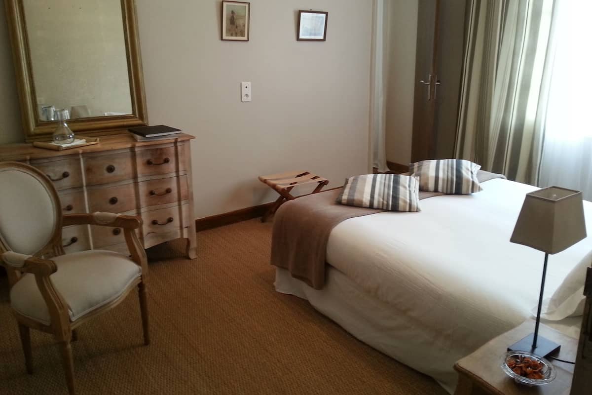 Una habitación de hotel con cama, sillón, cómoda, espejo y mesita de noche con lámpara.