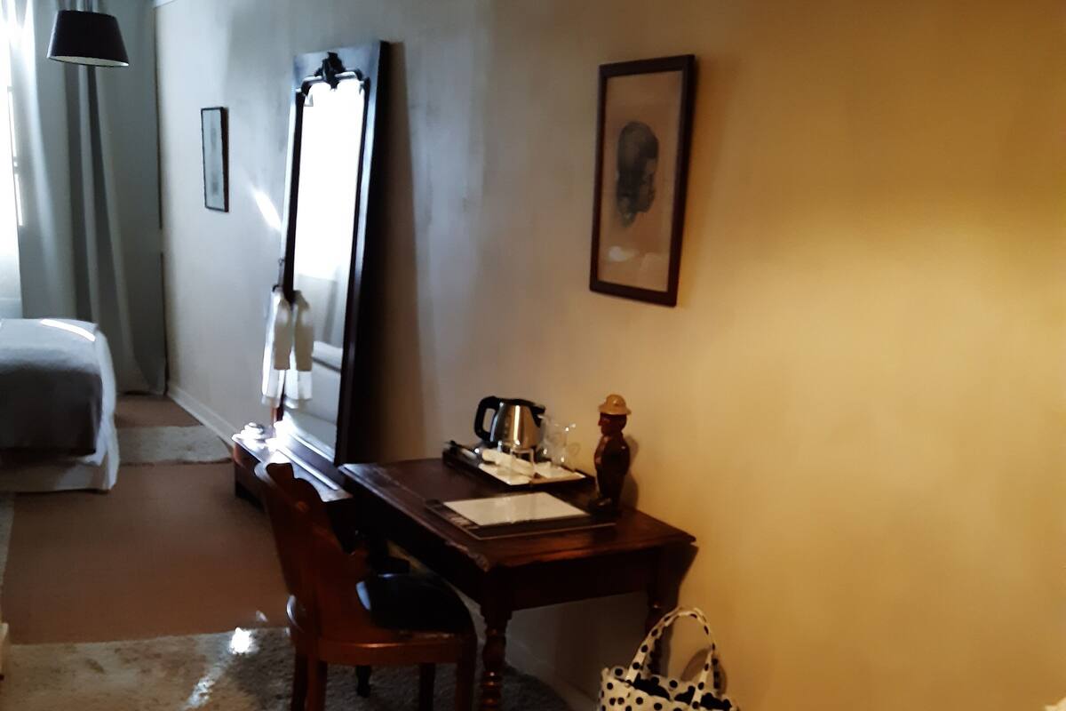 Una habitación de hotel con una cama, un escritorio con silla, una mesita con hervidor de agua, un bolso estampado con lunares y un taburete de madera.