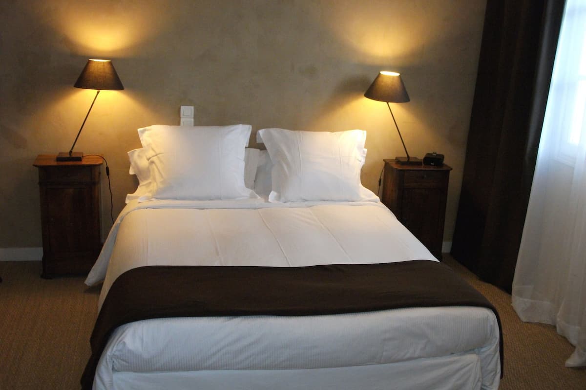 Una cama bien hecha con sábanas blancas, dos lámparas de noche y un plaid marrón oscuro.
