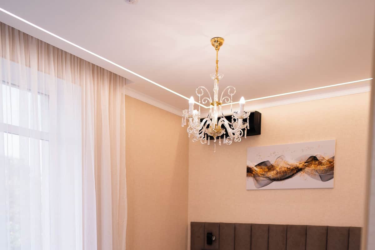 Quarto de hotel moderno com uma cama grande, um candelabro, uma mesa de cabeceira com abajur e vista para o exterior através de cortinas translúcidas.