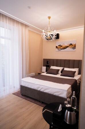 A modern hotel room with a large bed, a chandelier, a bedside table with a lamp, and a view of the outdoors through sheer curtains.