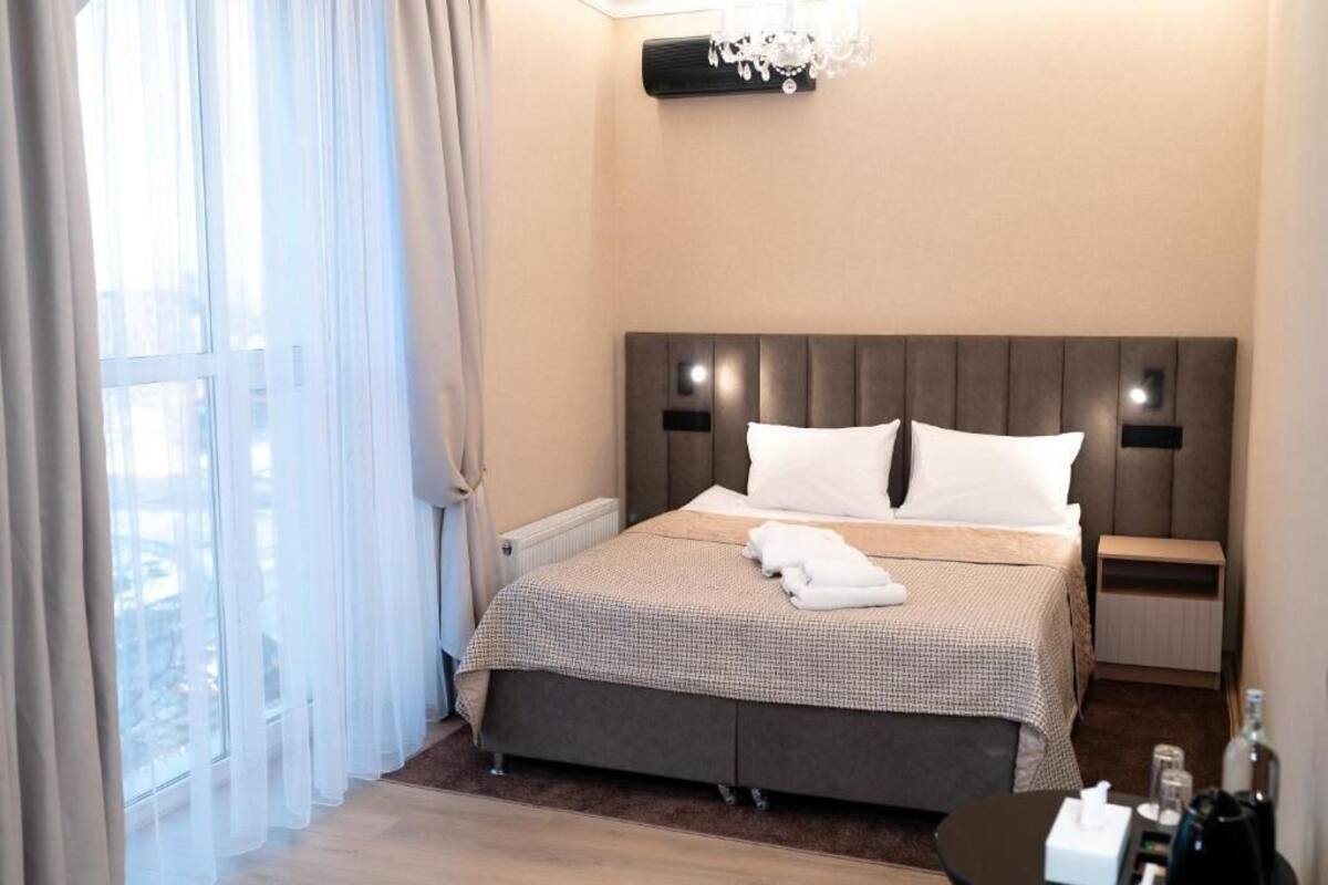 Quarto de hotel com uma cama grande, mesa de cabeceira, cortinas finas numa janela e um candelabro.