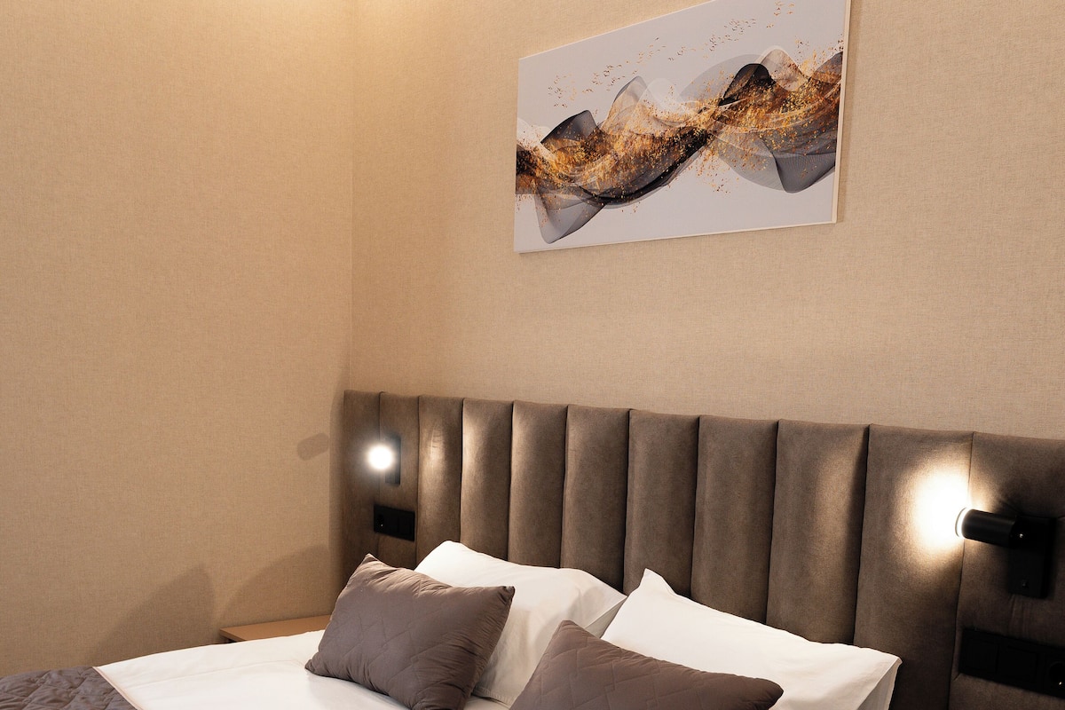 Uma cama bem arrumada com um edredom castanho, cabeceira com iluminação embutida e um quadro abstrato pendurado na parede.
