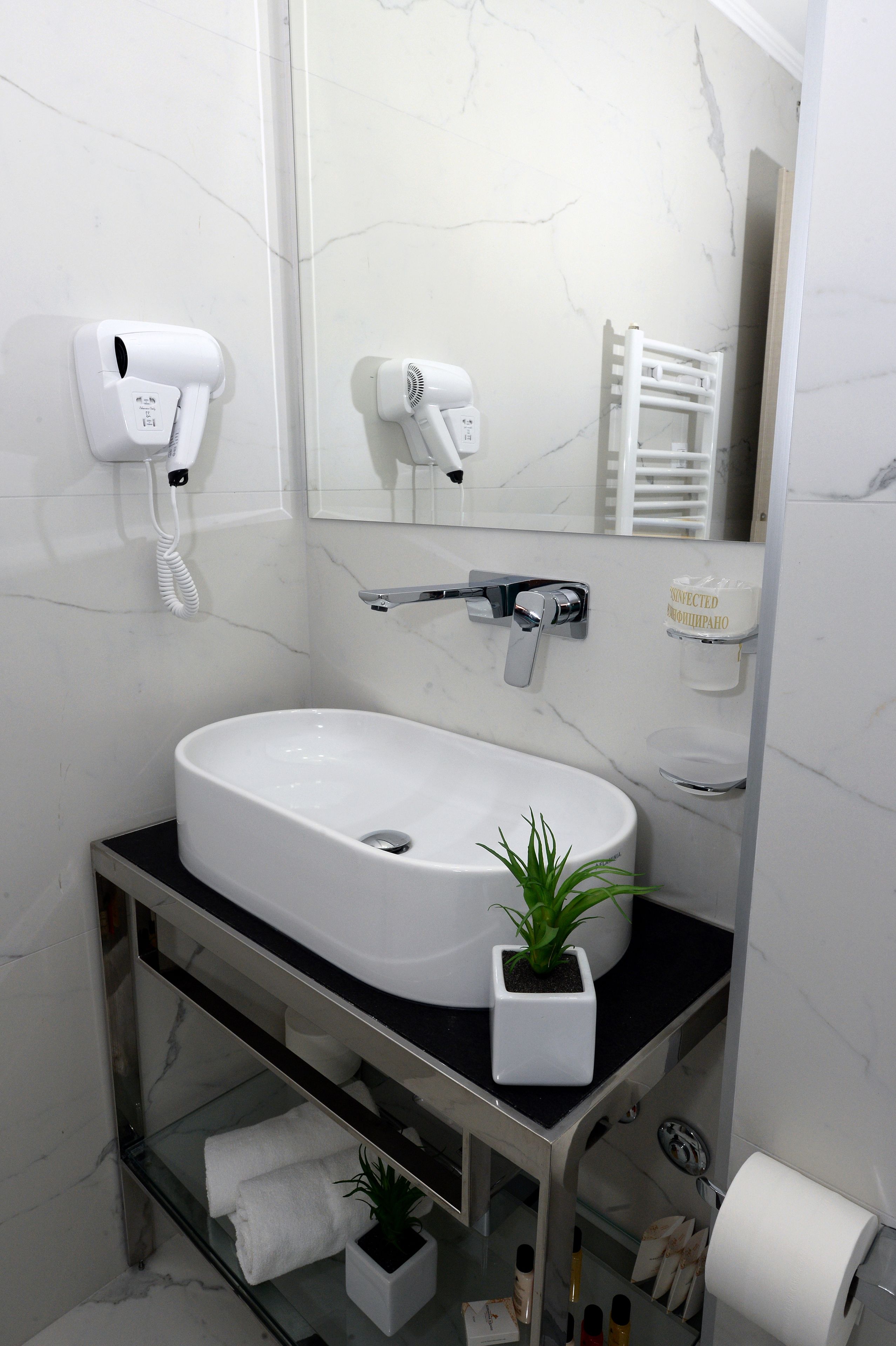 A bathroom with a white sink, two hair dryers mounted on the wall, a towel rack, a shelf with toiletries, and a mirror.