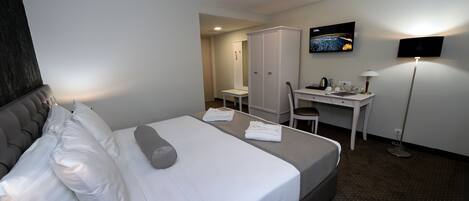 A hotel room with a bed, a desk, a chair, a wardrobe, a television, and a floor lamp.