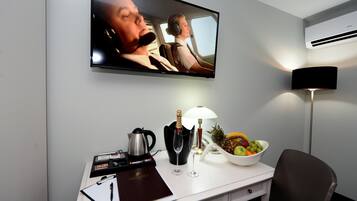 A hotel room with a flat-screen TV mounted on the wall, a desk with a laptop, a coffee maker, a bottle of champagne, a fruit bowl, and a lamp.