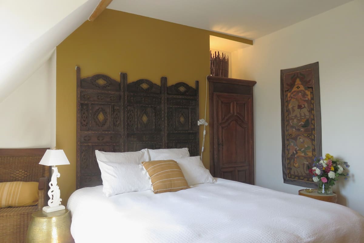 Un dormitorio con cama, mesita de noche, lámpara, cabecera de madera, tapiz en la pared y una silla de mimbre.