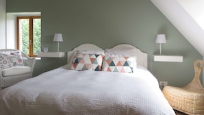 Un dormitorio con techo inclinado, cama con almohadones estampados, una silla de mimbre y una ventana con vistas a la vegetación.