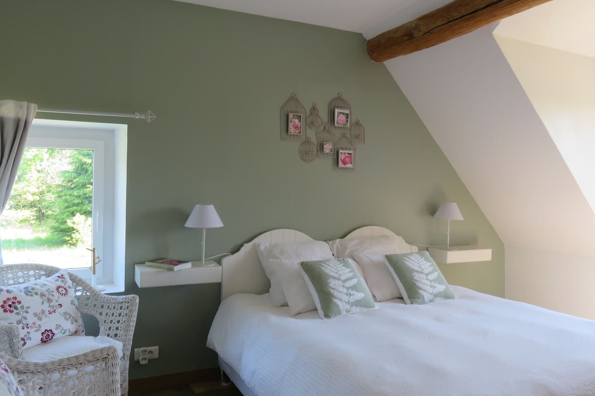 Un dormitorio con cama, dos lámparas de noche, una silla de mimbre, un ventanal y jaulas de pájaros en la pared.