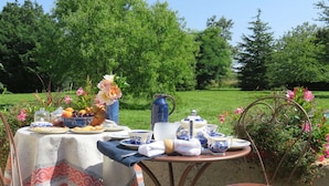 Mesa al aire libre con varios platos y una jarra, rodeada de follaje y flores.