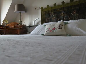 Un llit ben fet amb llençols blancs, un coixí decoratiu amb un disseny floral, una tauleta de nit de fusta amb una làmpada i un tapís de paret.