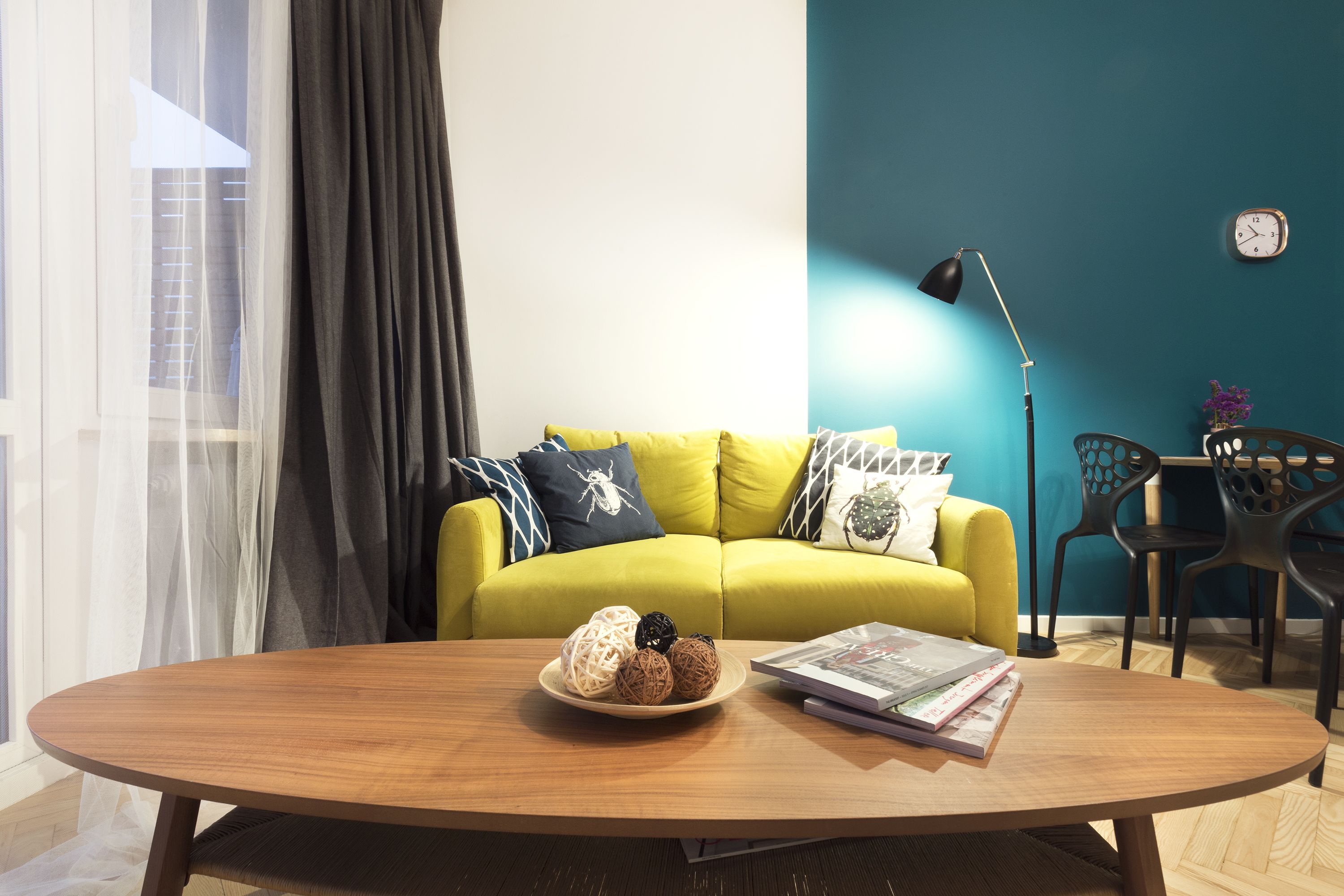 A modern living room with a yellow sofa, a round wooden coffee table with decorative balls and magazines, a floor lamp, and a view of the outside through sheer curtains.