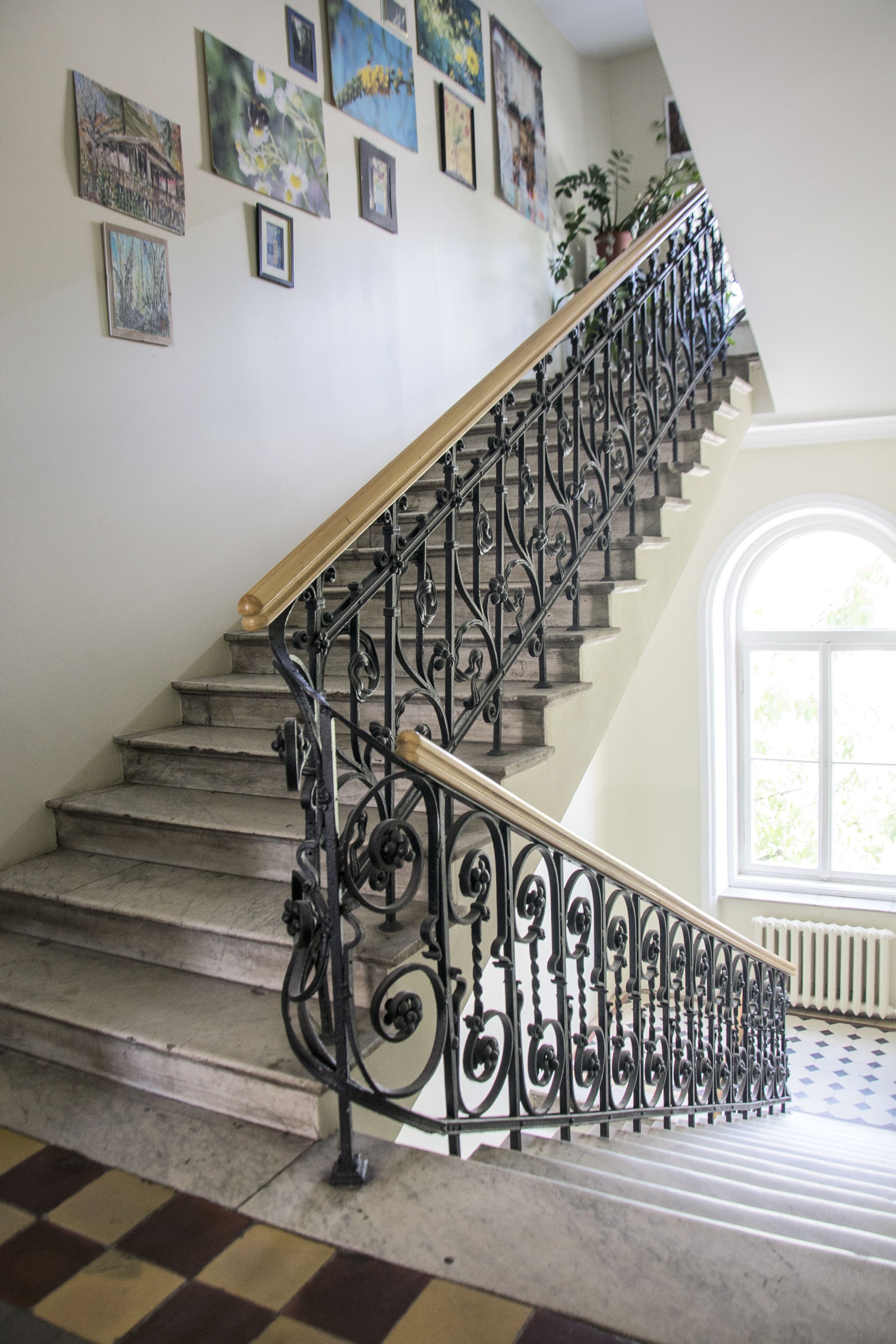 A staircase with ornate black iron railings and a wooden handrail, leading to a landing with a large arched window.