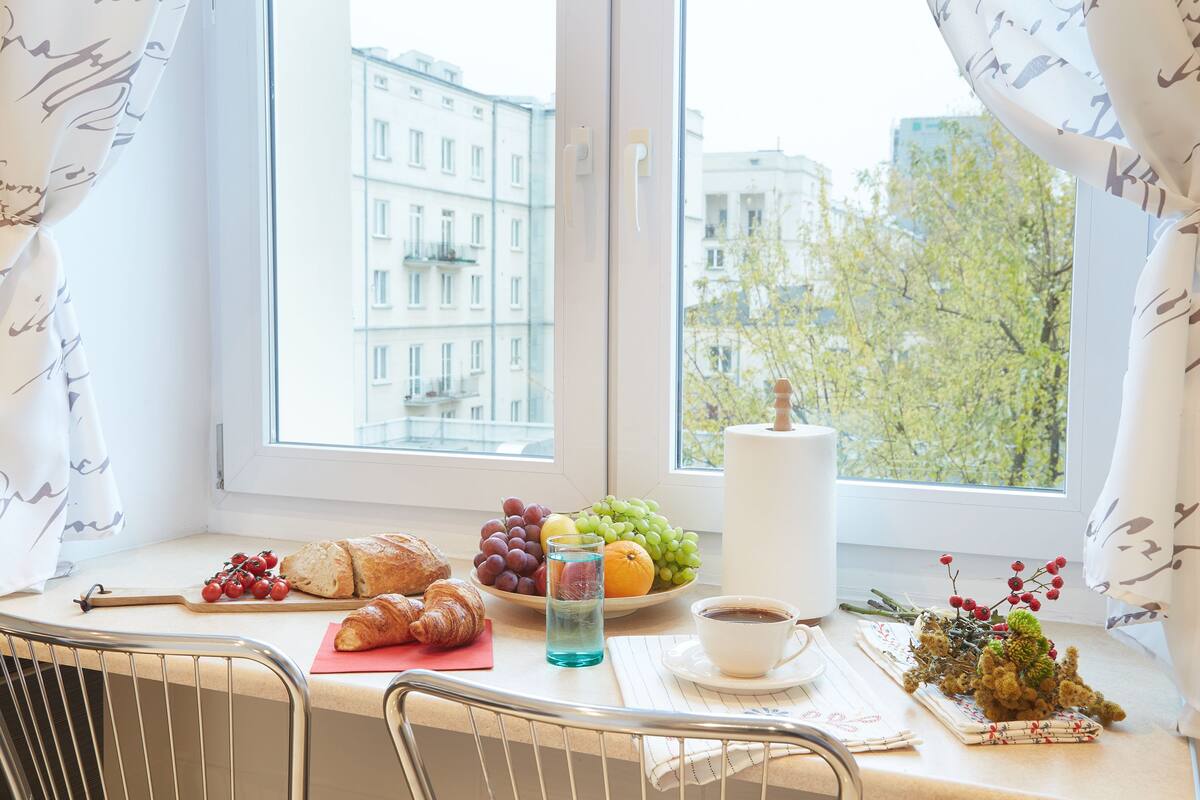 A breakfast table with croissants, grapes, an orange, and a cup of coffee by a window with a city view.
