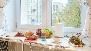 A breakfast table with croissants, grapes, an orange, and a cup of coffee by a window with a city view.
