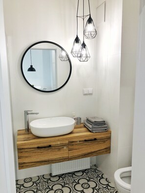 A bathroom with a round mirror, a white sink, wooden vanity, and a toilet. - Fox Center Apartments (Warsaw)