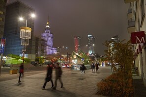 A nighttime cityscape with illuminated buildings, a prominent tower, and a pedestrian area with trees and a sign. - Fox Center Apartments (Warsaw)
