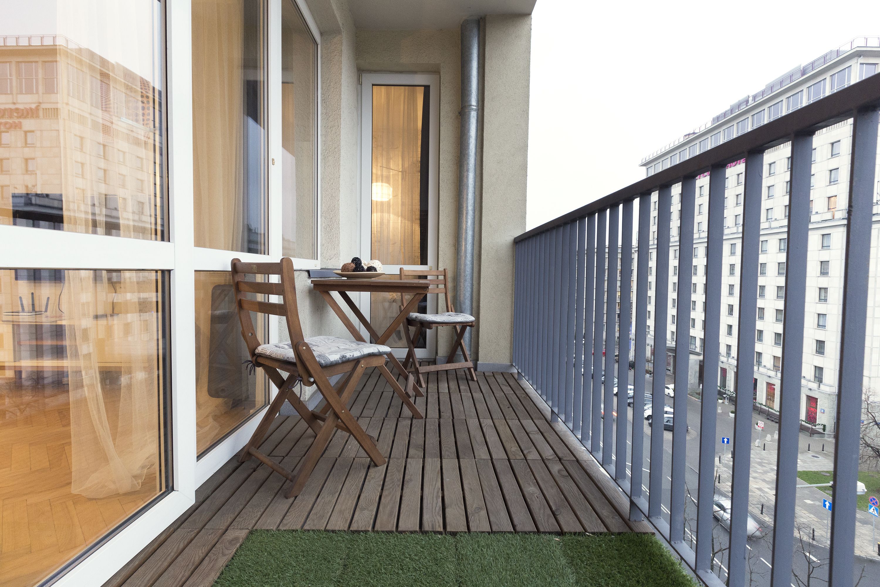 A balcony with wooden flooring, two wooden chairs, a small table, and a view of a cityscape through large windows.