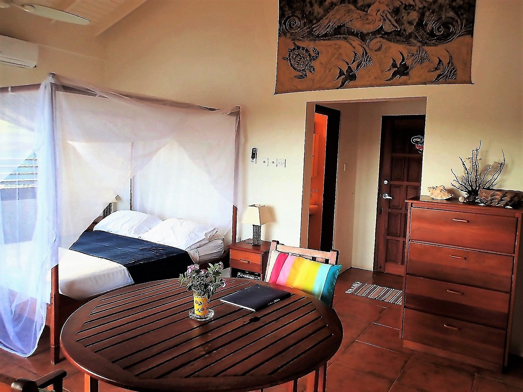 Una habitación con una cama, una mesa con un florero, un aparador y una puerta que da a otra habitación.