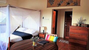 Una habitación con una cama, una mesa con un florero, un aparador y una puerta que da a otra habitación.