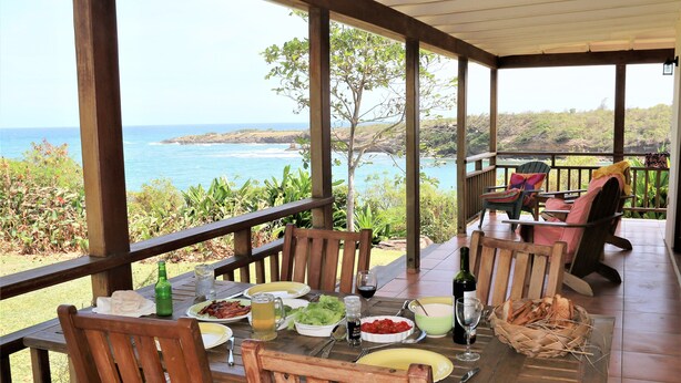 Uma área de refeições ao ar livre coberta, com móveis de madeira, uma mesa posta com comida e bebidas, e vista para o oceano.