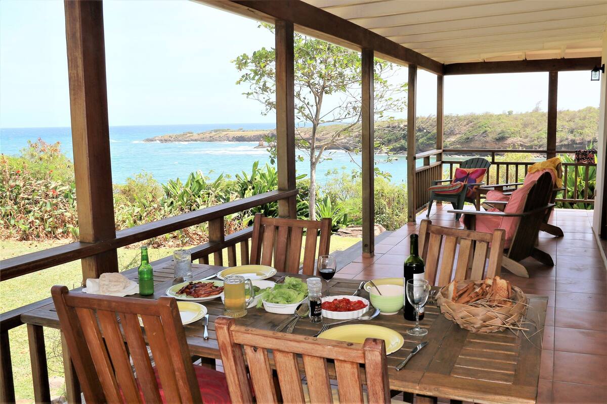 Een overdekt terras met houten meubels, een tafel gedekt met eten en drinken, en uitzicht op de oceaan.
