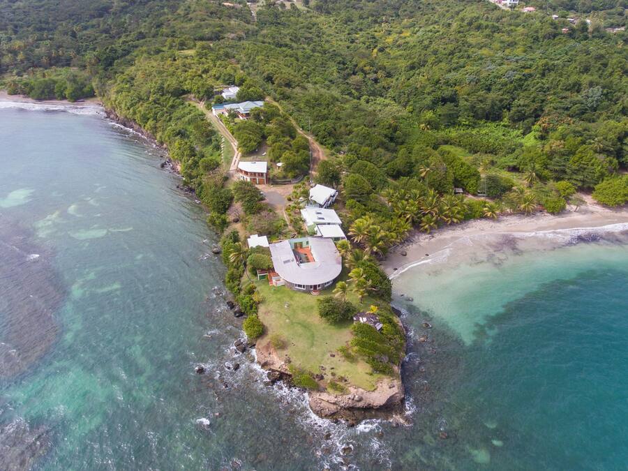 An aerial view of a coastal property with a large house, smaller structures, and lush greenery.