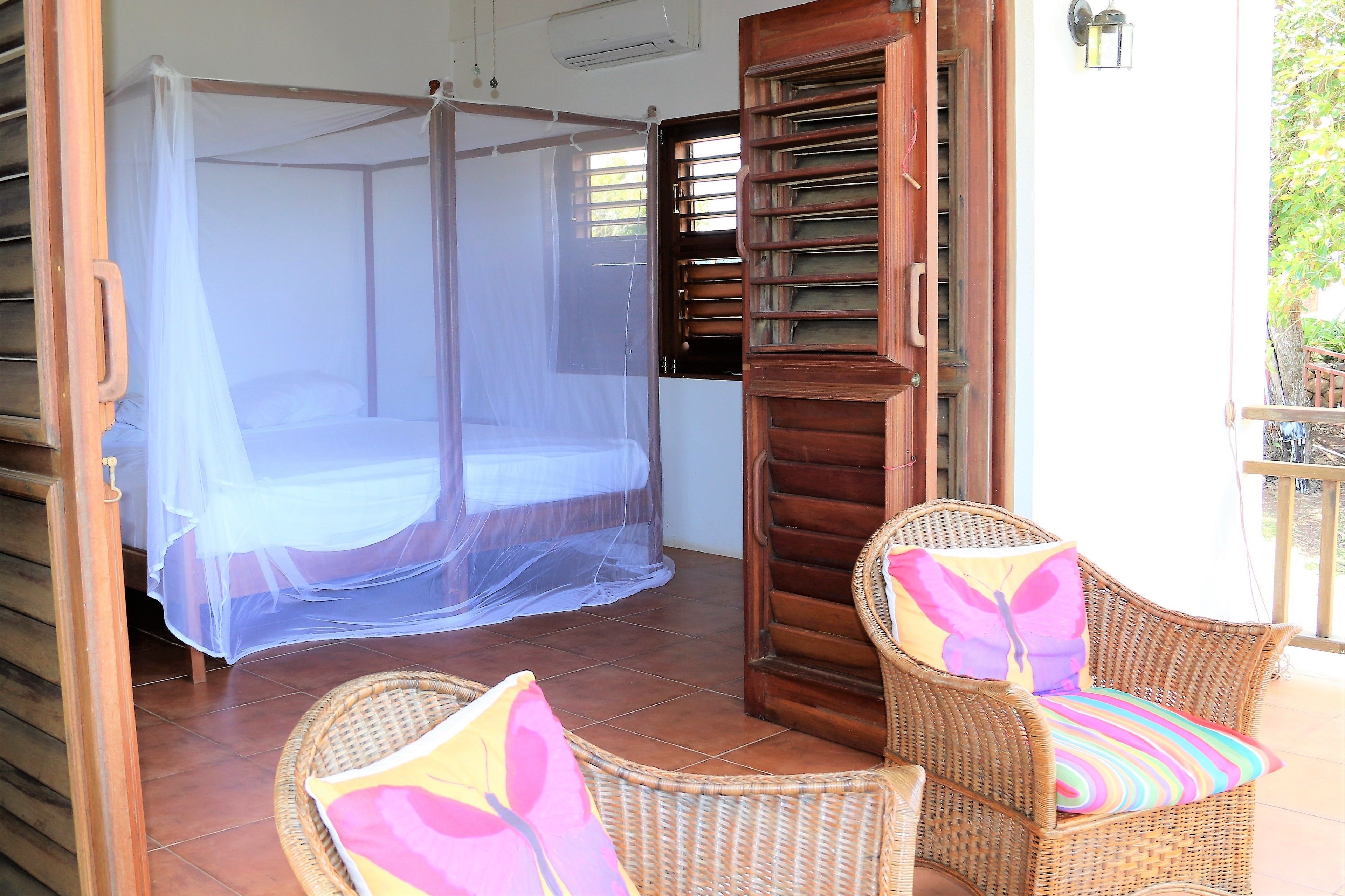 Una habitación con mosquitera sobre la cama, sillas de mimbre con cojines de colores y una puerta de madera con ventanal.
