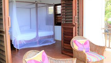 Una habitación con mosquitera sobre la cama, sillas de mimbre con cojines de colores y una puerta de madera con ventanal.
