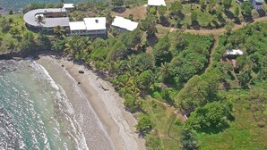 Vista aérea de uma propriedade costeira com praia, vários edifícios e vegetação exuberante.