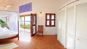 Um quarto com cama, uma cadeira, janela com persianas, ventoinha no teto e vista para o exterior.