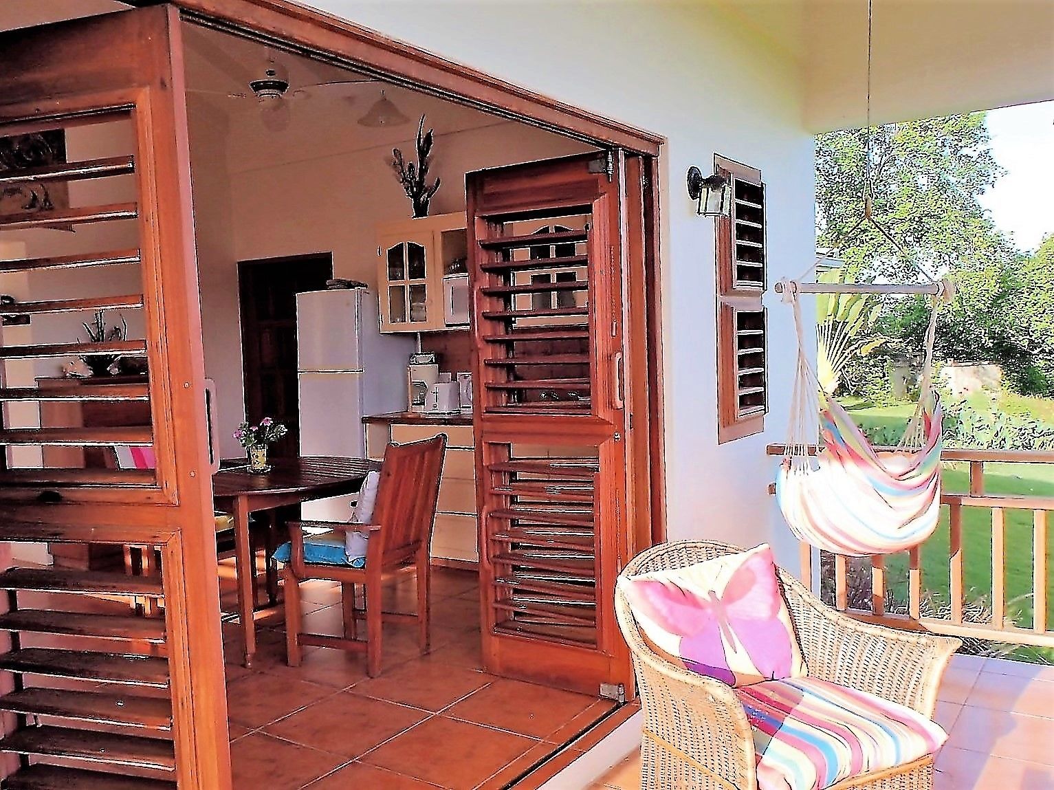 Une pièce avec un escalier en bois, un coin repas avec une table et des chaises, et un fauteuil en osier agrémenté d’un coussin coloré.