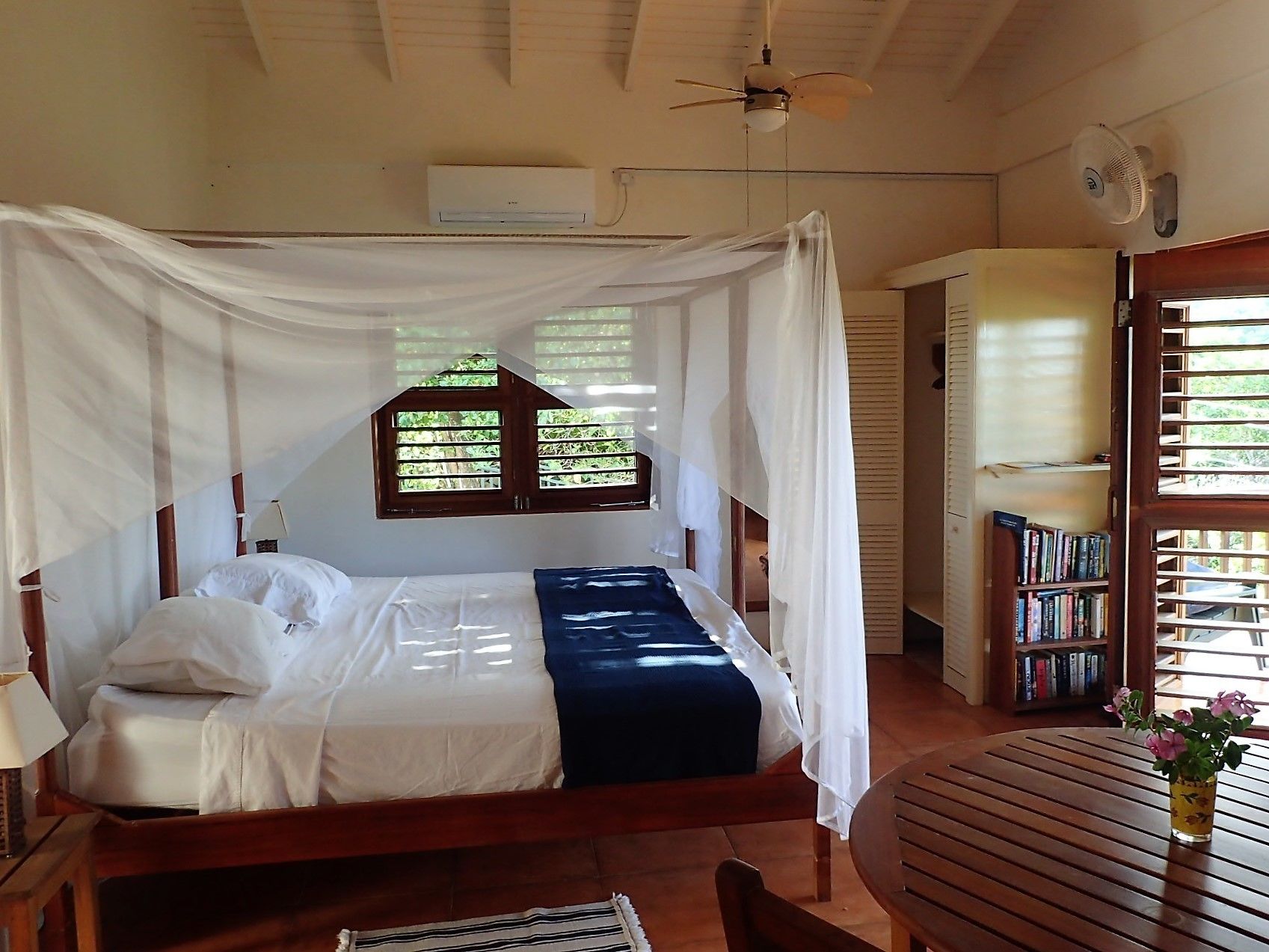 Una cama con dosel, una mesita con un florero y una estantería en una habitación con techo inclinado y ventana con contraventanas.