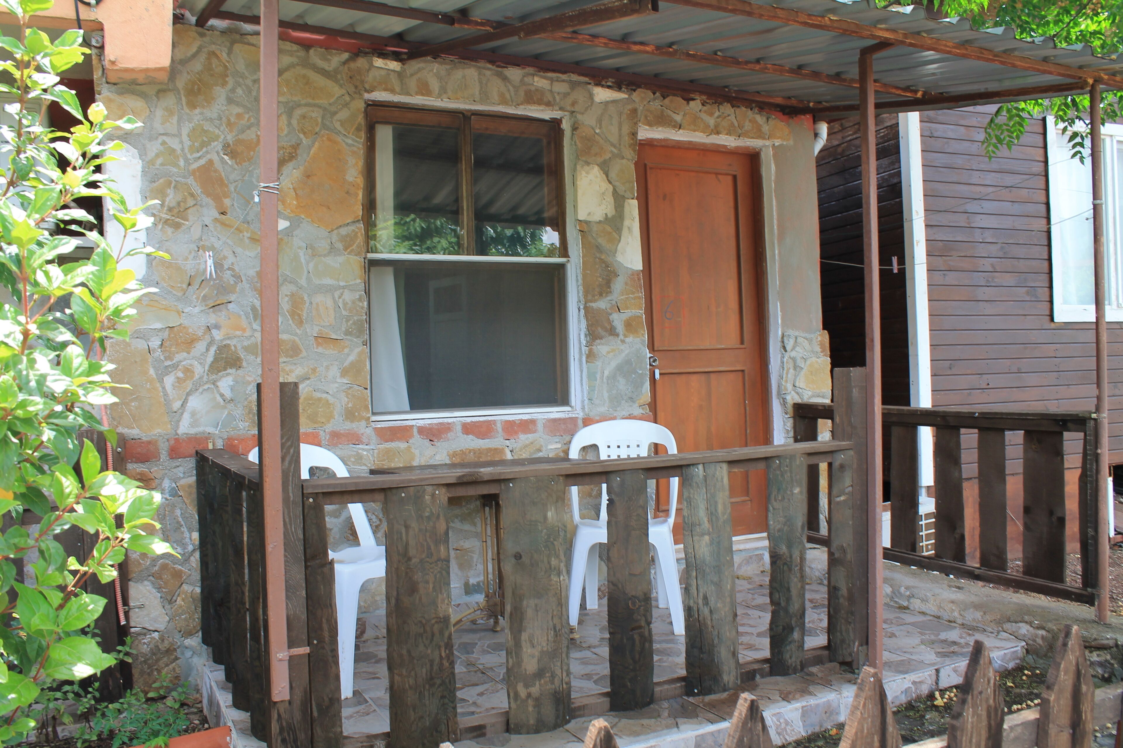 Un edificio de piedra con puerta y ventana de madera, un porche con barandillas de madera y una silla de plástico blanca.