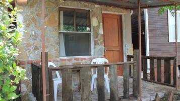 Un edificio de piedra con puerta y ventana de madera, un porche con barandillas de madera y una silla de plástico blanca.