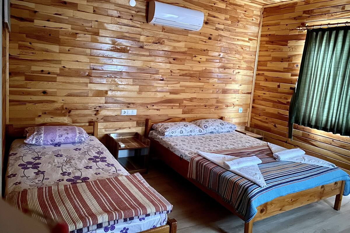 A wooden-paneled room with two beds, a nightstand, a wall-mounted air conditioner, and a window with curtains.